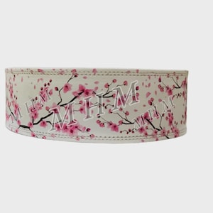 May include: A white weightlifting belt with a floral design of pink cherry blossoms and black branches. The belt has the letters "MHM" printed on it.