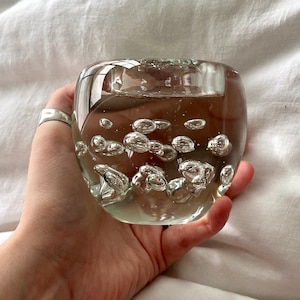 May include: A clear glass votive candle holder with bubble-like inclusions. The rounded, squat shape has a small opening at the top for a candle. The glass is held in a hand, resting on a white surface.