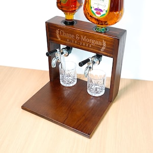 May include: A dark brown wooden liquor dispenser with two spouts and two glasses. The dispenser has a rectangular base and a top with two holes for bottles. The text "Chase & Morgan 5.15.2025" is engraved on the front.