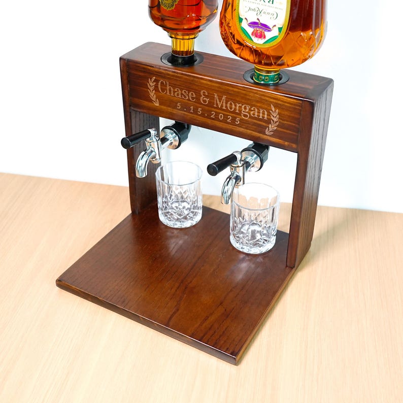 May include: A dark brown wooden liquor dispenser with two spouts and two glasses. The dispenser has a rectangular base and a top with two holes for bottles. The text "Chase & Morgan 5.15.2025" is engraved on the front.