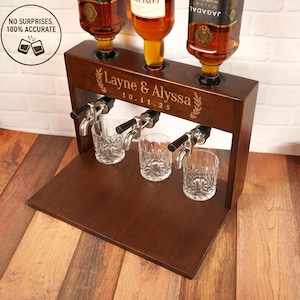 May include: A dark brown wooden liquor dispenser with three spouts and three glasses. The dispenser has the names "Layne & Alyssa" and the date "10.11.25" engraved on it. Three bottles are placed on top of the dispenser.