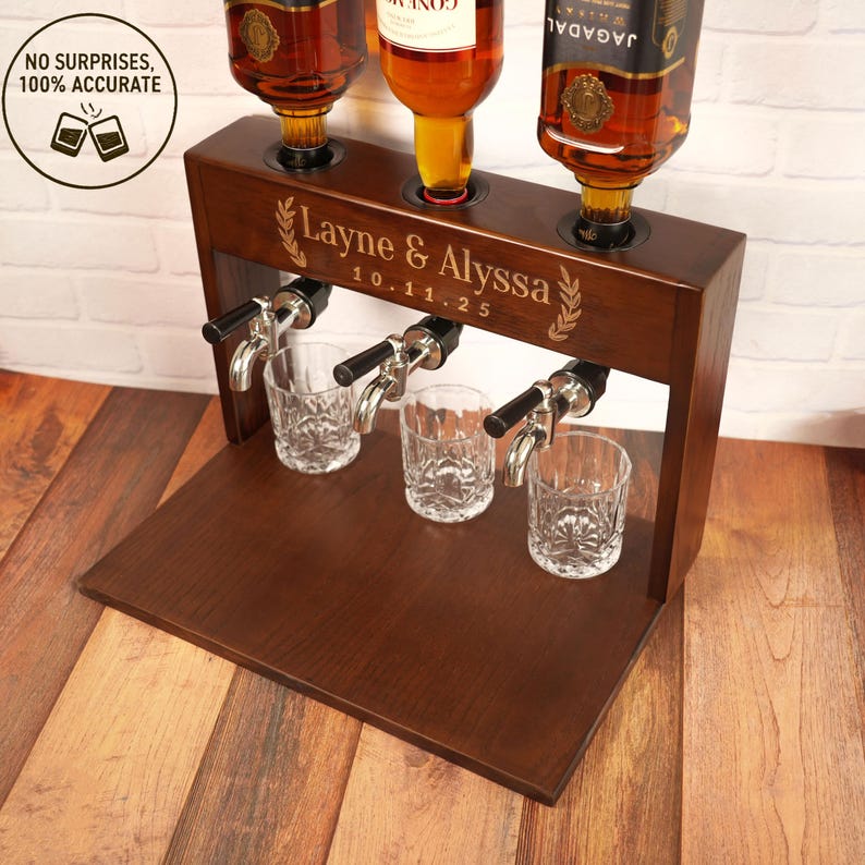 May include: A dark brown wooden liquor dispenser with three spouts and three glasses. The dispenser has the names "Layne & Alyssa" and the date "10.11.25" engraved on it. Three bottles are placed on top of the dispenser.
