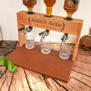 May include: A wooden liquor dispenser with three spouts and three crystal glasses. The dispenser is engraved with the names "Justin & Jackie" and the date "10.10.2008". The dispenser is made of light brown wood and has a dark brown base.