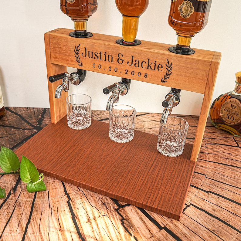 May include: A wooden liquor dispenser with three spouts and three crystal glasses. The dispenser is engraved with the names "Justin & Jackie" and the date "10.10.2008". The dispenser is made of light brown wood and has a dark brown base.