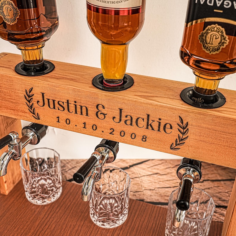 May include: A wooden liquor dispenser with three spouts, holding three bottles of amber-colored liquor. The wooden beam is engraved with "Justin & Jackie" and the date "10.10.2008". Two crystal glasses are positioned below the spouts.