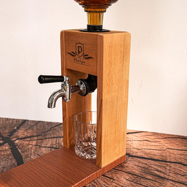 May include: Wooden liquor dispenser with a glass decanter and a chrome tap. The dispenser has a wooden frame with the name "Phelps" and a logo etched on the front. A clear, patterned glass sits below the tap.