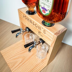 May include: Wooden liquor dispenser with two spouts and two glasses. The dispenser is engraved with "JOHNSON'S Bar". Two bottles are placed on top of the dispenser. The spouts are made of metal with black handles. The glasses are clear and have a patterned design.