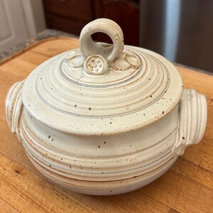 May include: A round, off-white ceramic lidded casserole dish with brown speckles and horizontal ridges. The lid features a central loop handle decorated with small, raised circular floral motifs. Two side handles are integrated into the body.