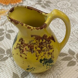 May include: A small yellow ceramic pitcher featuring a curved handle and a pouring spout. The exterior surface is decorated with brown mottled accents and dark green patches, with the text The Hen House printed in a cursive script on the front.