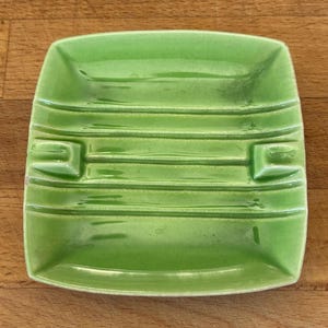 May include: A square, light green ceramic ashtray with a glossy finish. The piece features a series of horizontal, raised ridges across the center and two distinct notches on opposite sides for resting items, all within a shallow basin.