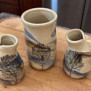 May include: Three ceramic vessels with a glossy cream-colored glaze, featuring hand-painted black tree silhouettes and blue landscape accents. Two smaller vessels include side spouts with white wicks, while the central vessel is a tall cylinder.