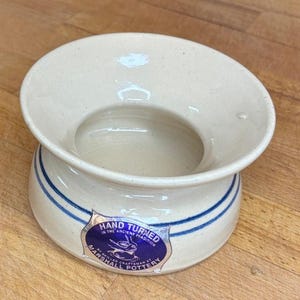 Puede incluir: Un cuenco de cerámica de color crema con un borde ancho y acampanado, y dos finas rayas azules horizontales alrededor de la base. Una pegatina azul y plateada en el lateral dice: HAND TURNED IN THE ANCIENT TRADITION BY SKILLED CRAFTSMAN AT MARSHALL POTTERY.