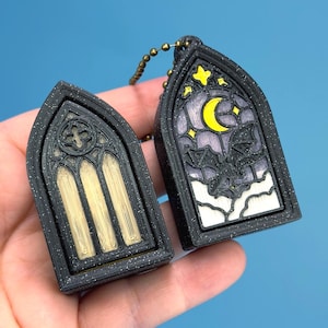 May include: Two black gothic window-shaped keyrings. One features a cross design, the other a moon, stars, and a bat silhouette. The keyrings have gold and white accents and are connected by a beaded chain.