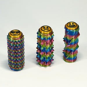 May include: Three colorful, cylindrical objects with textured surfaces and emoji faces on top. The objects are primarily rainbow-colored with gold accents. The emoji faces include a smiling face with sunglasses, a sad face, and a smiling face.
