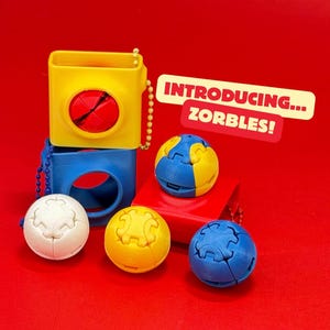 May include: A collection of colorful, geometric puzzle toys, including yellow, blue, and white spheres and cube-shaped containers. The text "INTRODUCING... ZORBLES!" is displayed on a red background.