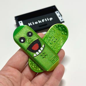 May include: A green toy skateboard with a cartoon pickle face design. The skateboard has a smiling face with white teeth, black eyes, and a red mouth. The text "Kickflip" is on a black box in the background.