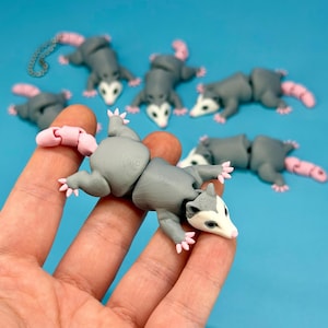 May include: Articulated grey and pink opossum figurine with a white face and pink feet. The opossum is lying on its belly. The background is a solid light blue.