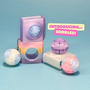 May include: A collection of colourful, geometric objects against a blue backdrop. Two stacked, lavender-coloured, square-shaped items with a circular design are linked by a chain. The text "INTRODUCING... ZORBLES!" is displayed. Spherical objects in pink, blue, and purple are also present.