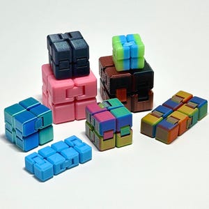 Può includere: Una collezione di colorati cubi e giocattoli fidget. I cubi sono in vari colori, tra cui blu, rosa, verde e marrone. Alcuni sono assemblati in forme di cubo, mentre altri sono in configurazioni lineari. Lo sfondo è bianco.