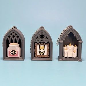 May include: Three grey arched display charms with silver ball chains. The first contains a pink potion bottle with a moon symbol. The second features a framed picture of two yellow butterflies. The third holds a cluster of white pillar candles.