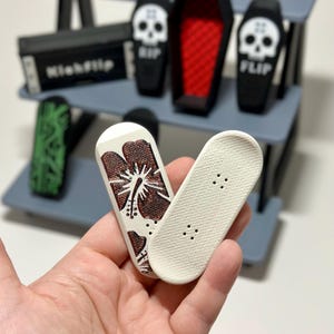 May include: Miniature finger skateboards, one with a brown floral design and the other plain. The set includes a gray shelf with miniature coffins, a box labeled "Kickflip", and a green skateboard.