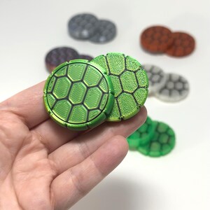 May include: Two circular tokens with a green turtle shell pattern featuring black hexagonal outlines. One token has a smooth, lined surface texture, while the other has a granular, textured finish. Additional tokens in various colours are visible.