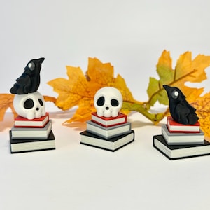 May include: Three miniature Halloween sculptures. Each features a white skull or black crow atop a stack of books. The books are black, grey, and red. Autumn leaves are in the background. A spooky and decorative item.