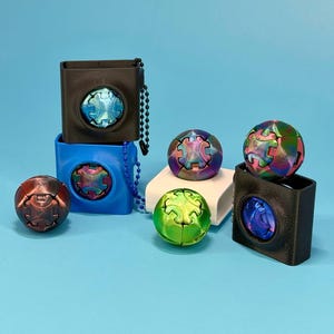 May include: A collection of colorful, spherical fidget toys and square containers with circular cutouts. The fidget toys have a gear-like design and iridescent finishes. The containers are black and blue, with a chain attached to the black container.