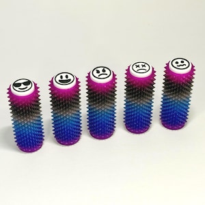 May include: Five spiky cylinders with emoji faces. The cylinders are a gradient of purple, blue, black and pink. Each cylinder has a different emoji face, including a smiling face, a face with sunglasses, and a sad face.
