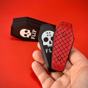 May include: Black coffin-shaped cardholder with a white skull graphic and the word "FLIP". A red and black coffin-shaped skateboard is held in a hand. The background is a solid red.