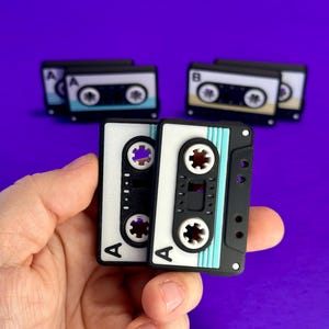 May include: Two miniature cassette tapes, one black and one white, are held in a hand against a purple background. The white tape has a teal stripe. Other tapes are visible in the background. The tapes have the letter "A" printed on them.
