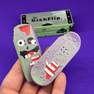 May include: Two small, textured fingerboards with zombie-themed graphics in shades of green, purple, and red. One board features a face with a wide mouth, while the other has a textured surface. A green box labelled Kickflip is visible in the back.