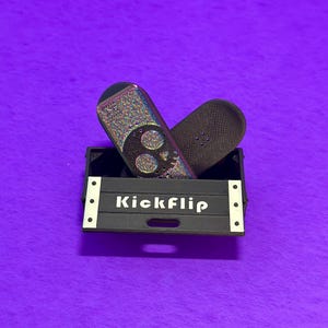 May include: Two miniature skateboards rest in a black box with the word "Kickflip" in white. One skateboard has a skull design with iridescent glitter, and the other is dark brown. The box has white accents and sits on a purple surface.