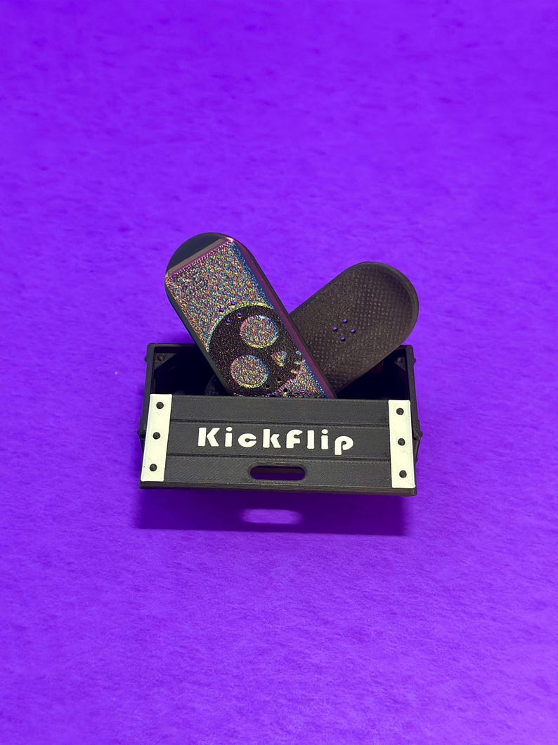 May include: Two miniature skateboards rest in a black box with the word "Kickflip" in white. One skateboard has a skull design with iridescent glitter, and the other is dark brown. The box has white accents and sits on a purple surface.