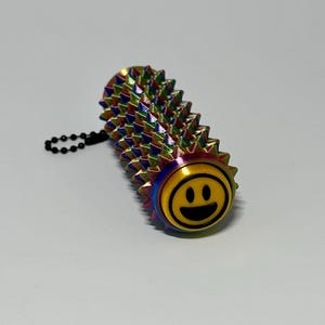 Puede incluir: Un juguete antiestr&eacute;s de metal multicolor y puntiagudo con una carita sonriente en un extremo. El juguete tiene forma cil&iacute;ndrica con una cadena adjunta. Los colores incluyen tonos de azul, verde, amarillo y morado.