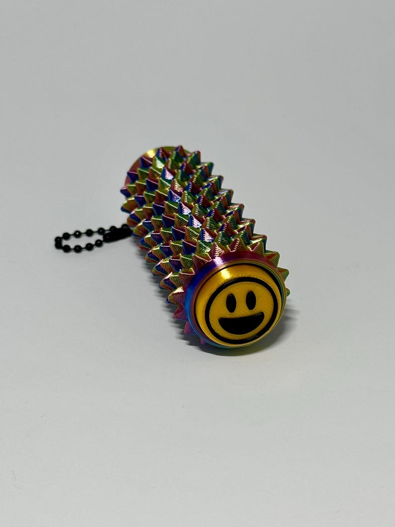 Puede incluir: Un juguete antiestr&eacute;s de metal multicolor y puntiagudo con una carita sonriente en un extremo. El juguete tiene forma cil&iacute;ndrica con una cadena adjunta. Los colores incluyen tonos de azul, verde, amarillo y morado.