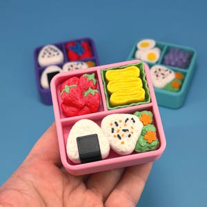 May include: Miniature bento boxes with various food items. The pink box contains strawberries, rice balls, and yellow omelet. Other boxes in purple and teal have similar food items. The food items are white, red, yellow, and green.
