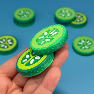 May include: Two circular, green, textured cucumber slice replicas with white seed details. The slices feature a layered, ridged surface texture on the top and a bumpy, realistic rind pattern on the sides, with additional similar slices scattered nearby.