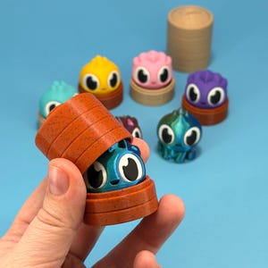 May include: An orange container is held, revealing a blue creature with large eyes. Other colorful creatures with large eyes sit on small wooden platforms. The background is a solid light blue.