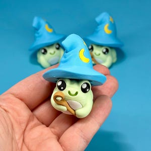 May include: A light green frog figurine wearing a blue wizard hat with a yellow crescent moon. The frog holds a small brown wand. Two other figurines are slightly out of focus in the background. The background is a solid light blue.
