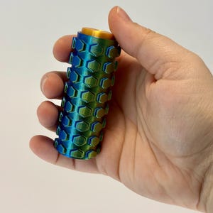 Puede incluir: Un objeto cil&iacute;ndrico con un patr&oacute;n de panal en tonos azules y verdes, coronado con una tapa dorada. El objeto se sostiene en una mano sobre un fondo blanco. El dise&ntilde;o presenta un patr&oacute;n hexagonal repetitivo.