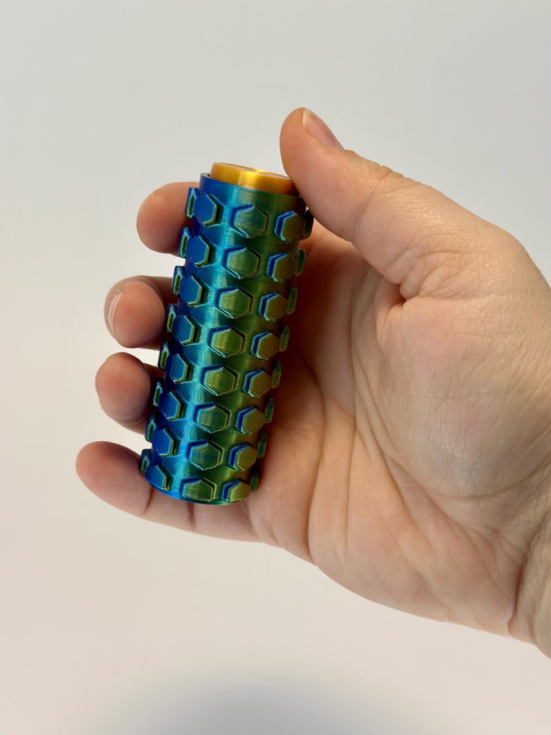 Puede incluir: Un objeto cil&iacute;ndrico con un patr&oacute;n de panal en tonos azules y verdes, coronado con una tapa dorada. El objeto se sostiene en una mano sobre un fondo blanco. El dise&ntilde;o presenta un patr&oacute;n hexagonal repetitivo.