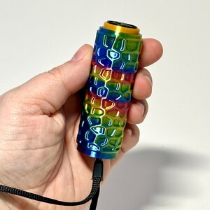 Puede incluir: Un objeto cil&iacute;ndrico con un exterior de colores del arco&iacute;ris y un patr&oacute;n de panal. La parte superior es dorada y una correa negra est&aacute; unida en la parte inferior. El objeto se sostiene en una mano sobre un fondo blanco.