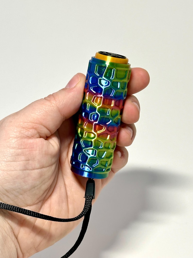 Puede incluir: Un objeto cil&iacute;ndrico con un exterior de colores del arco&iacute;ris y un patr&oacute;n de panal. La parte superior es dorada y una correa negra est&aacute; unida en la parte inferior. El objeto se sostiene en una mano sobre un fondo blanco.