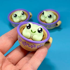 May include: Three small, light green frog figurines with large black eyes and smiling mouths, each sitting inside a tan teacup. The cups feature a purple rim and a purple leaf pattern around the exterior. The figurines are made of a smooth material.