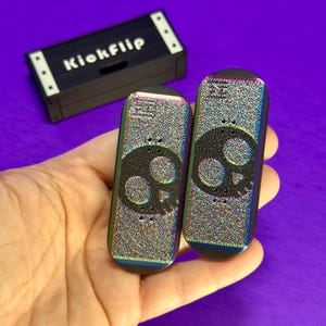 May include: Two finger skateboards with a skull design and a black carrying case that says "Kickflip". The skateboards have a colorful, iridescent finish. The carrying case is black with white accents.