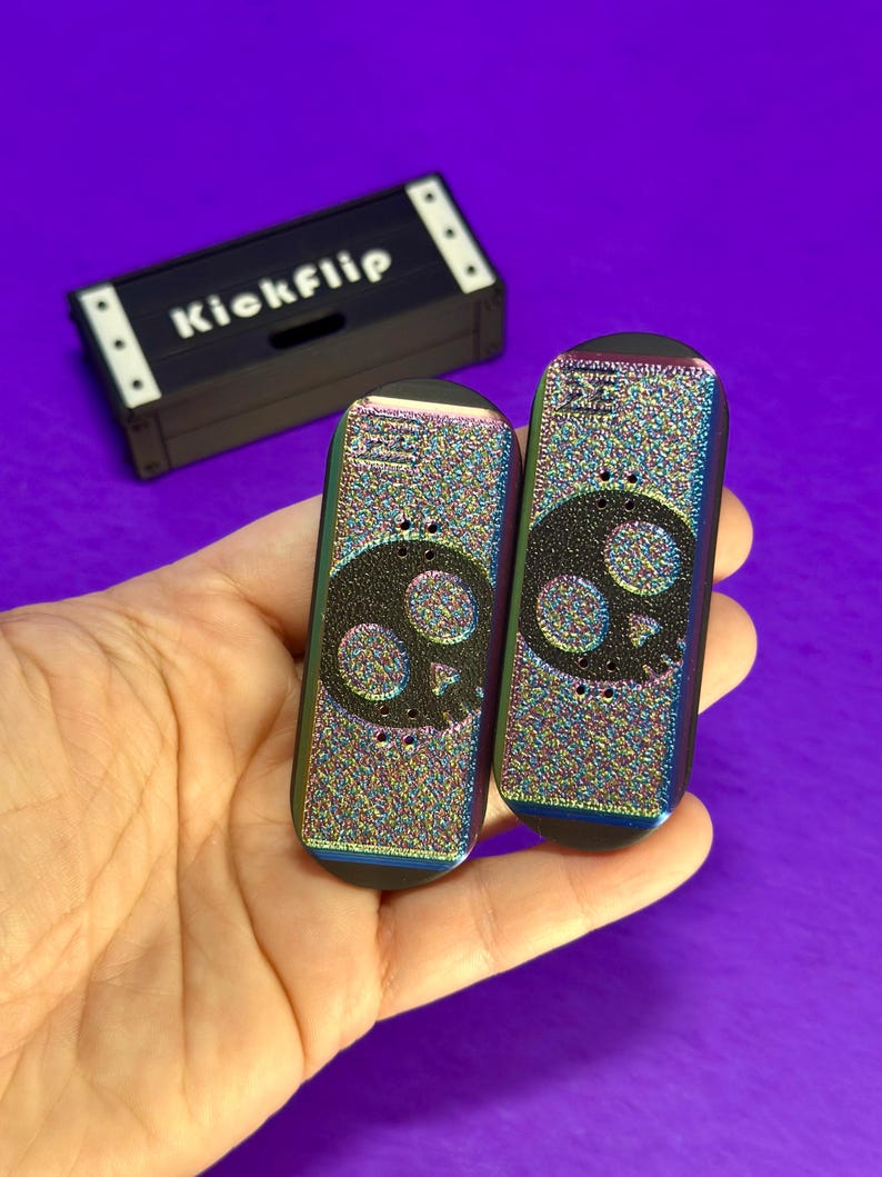 May include: Two finger skateboards with a skull design and a black carrying case that says "Kickflip". The skateboards have a colorful, iridescent finish. The carrying case is black with white accents.