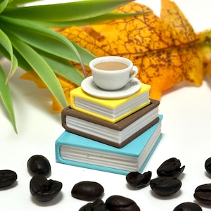 May include: A small white coffee cup and saucer rest on a stack of three miniature books in blue, brown, and yellow. Dark brown coffee beans are scattered around the books. A green plant and a yellow leaf are in the background.