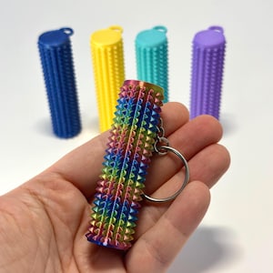 May include: A hand holds a rainbow-coloured, textured keychain with a silver key ring. In the background are four cylindrical objects in blue, yellow, teal, and purple. The objects have a similar textured design.