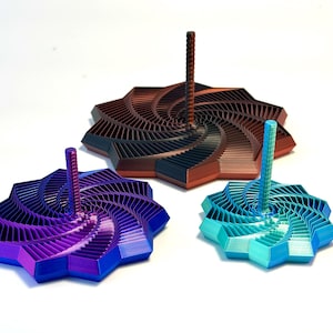 May include: Three decorative, geometric tops in shades of purple, teal, and copper. Each top features a spiral design and a central, cylindrical post. The tops are arranged on a white surface, showcasing their intricate, layered construction.